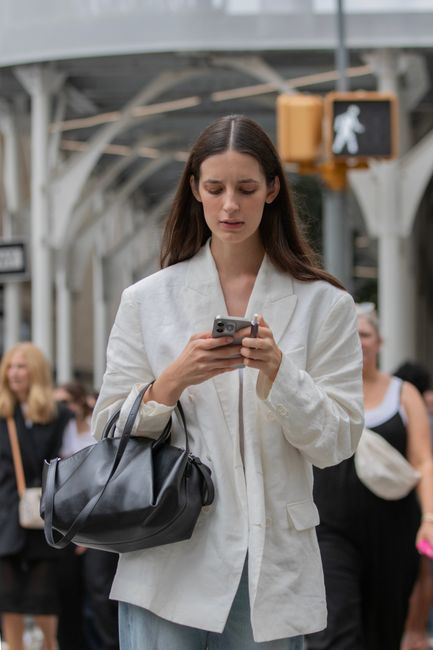 A young woman with long dark hair, dressed in a cream-colored blazer and light blue jeans, stands outdoors in a bustling urban environment, possibly a market or train station, with a partly cloudy sky