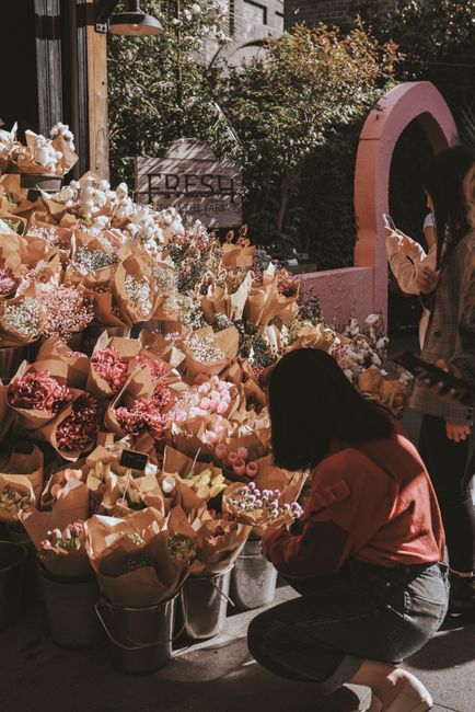 A market stall displays an extensive selection of fresh flowers arranged in buckets and wrapped in brown craft paper. The bouquet contains various flower types including pink, white, and purple blooms