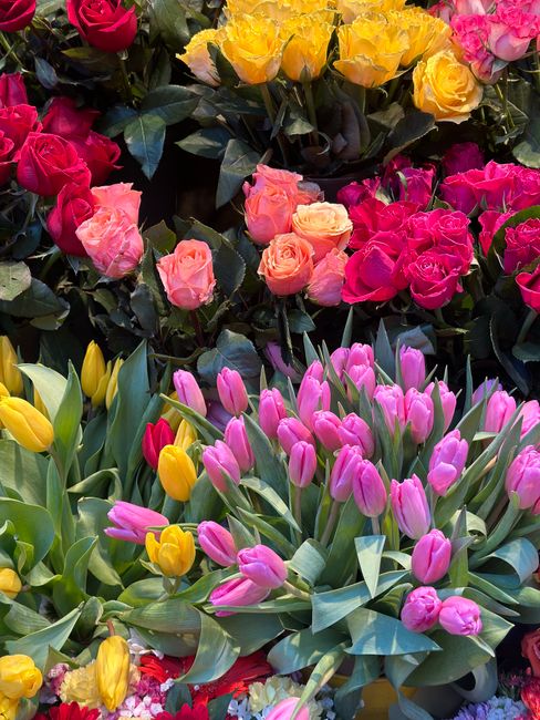 A vibrant display of fresh flowers arranged in a floral shop setting, featuring a mix of pink tulips with slender green stems and elongated leaves, bright yellow tulips with smooth petals, and cluster