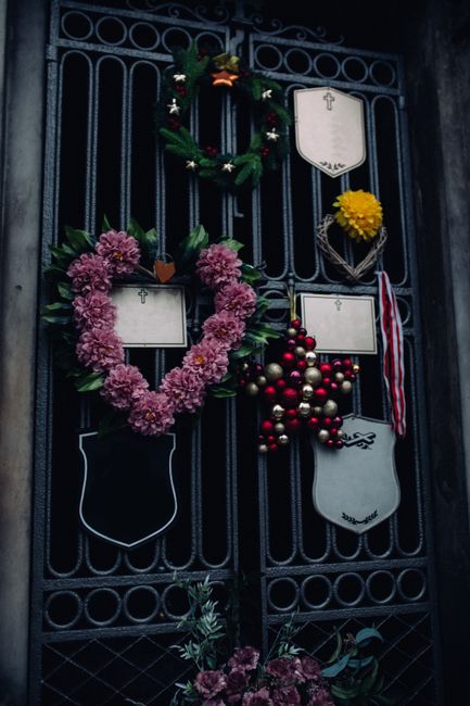 A decorative black wrought iron gate adorned with various floral and festive arrangements. On the left, there is a large heart-shaped wreath made of pink chrysanthemums with green foliage, hanging alo