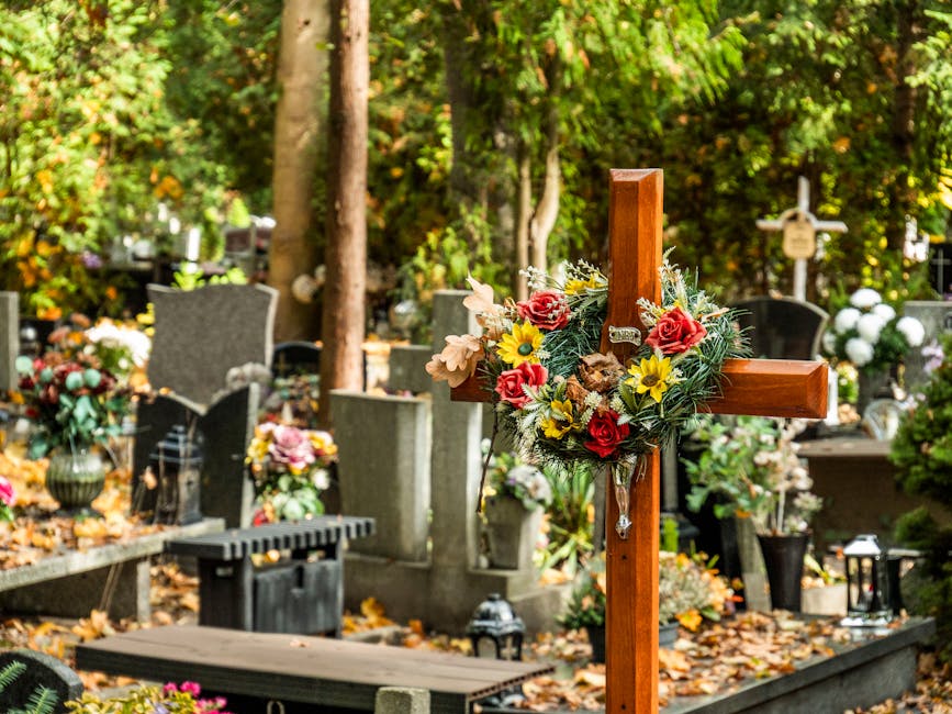 A wooden cross decorated with a vibrant floral arrangement placed at a cemetery site. The arrangement includes red roses, yellow daisies, light-colored lilies, and greenery, all fresh and petal-rich. 