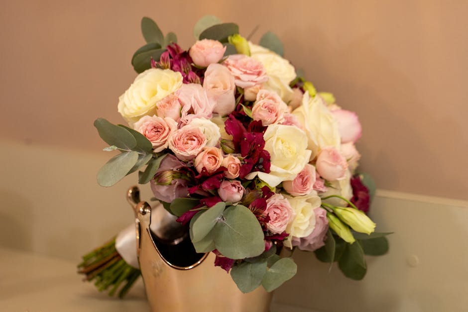 A floral bouquet featuring a mix of fresh flowers including pale pink roses, creamy white roses, and deep burgundy blooms, arranged with eucalyptus leaves and other greenery. The flowers are tightly c