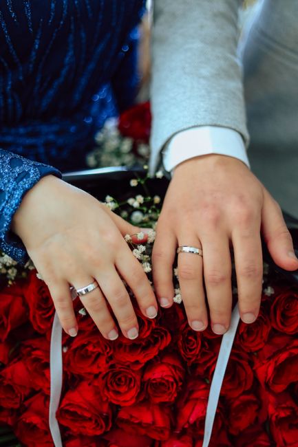 Close-up image capturing the hands of a newlywed couple resting on a lush, densely packed arrangement of vibrant red roses with velvety petals. The bride's hand displays a silver wedding band, and she