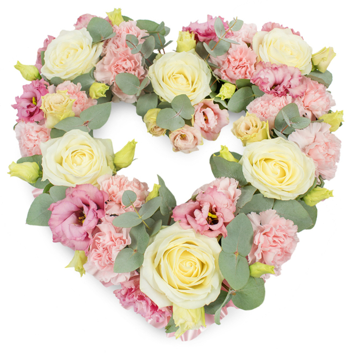 A beautifully composed heart-shaped floral tribute is displayed against a bright white background, its pastel colours and detailed textures drawing the eye into every corner of the design. The heart is full and well-rounded, with no harsh edges, as though it has been patiently built up layer by layer. Cream and pale yellow roses form the main structure; each rose is at an ideal stage of opening, with tightly cupped centres and outer petals just beginning to curve back in soft, velvety spirals. Filling the spaces between them are clusters of light pink carnations, their countless ruffled petals pressing close together to create a plush, almost quilted effect. Deeper rose-pink and dusky mauve lisianthus blooms are scattered throughout, their looser, gently fluted petals adding a romantic, slightly more dramatic note that still remains soothing. Silvery-green eucalyptus leaves weave naturally between the blossoms, both along the outer edge of the heart and in its inner curve, giving a subtle definition to the shape and hinting at a cool, fresh scent. Here and there, small yellow-green buds peep out from the mass of petals, adding flickers of life and showing that the stems are newly cut. The palette of creams, soft pinks, gentle yellows, and muted greens creates an overall impression of tranquillity and compassion, making this an ideal wreath for funeral services, memorials, or anniversaries around New Cross and the surrounding streets near Deptford and Brockley. It feels like a tribute carefully prepared by a local florist who understands the area and the significance of saying farewell with something truly heartfelt and beautiful.